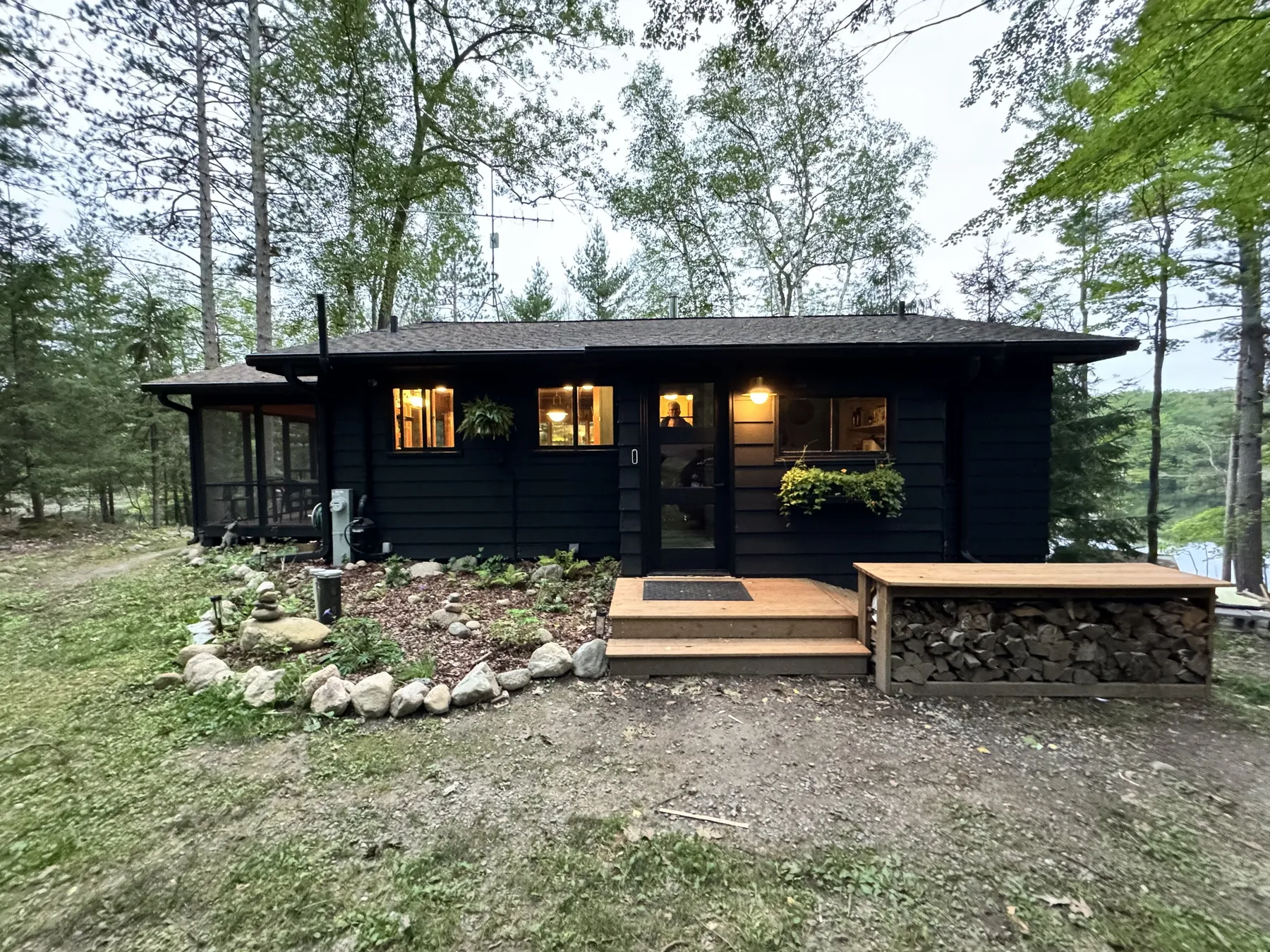 The cabin after the remodel, dark siding and lit windows at dusk, with new steps and a firewood bench at the front door